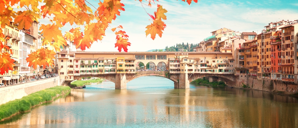 A covered foot bridge crossing a canal in Florence, Italy. A tree with Fall colored leaves hangs in the foreground.