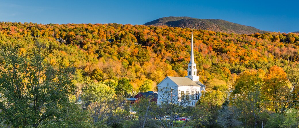 A historic looking church in Stowe, Vermont. Surrounding the church is a hillside of trees with Fall colored leaves.