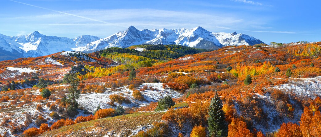 View from a mountaintop of other snow covered mountains near Aspen, Colorado. A nearby hillside is covered with trees with orange colored leaves in the Fall.