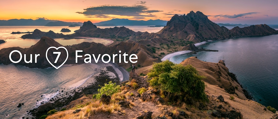 Aerial view of Komodo National Park in Indonesia. There is a penninsula extending out into the ocean. A man is standing in the distance taking a photograph. There is a title overlay that reads: Our 7 Favorite.