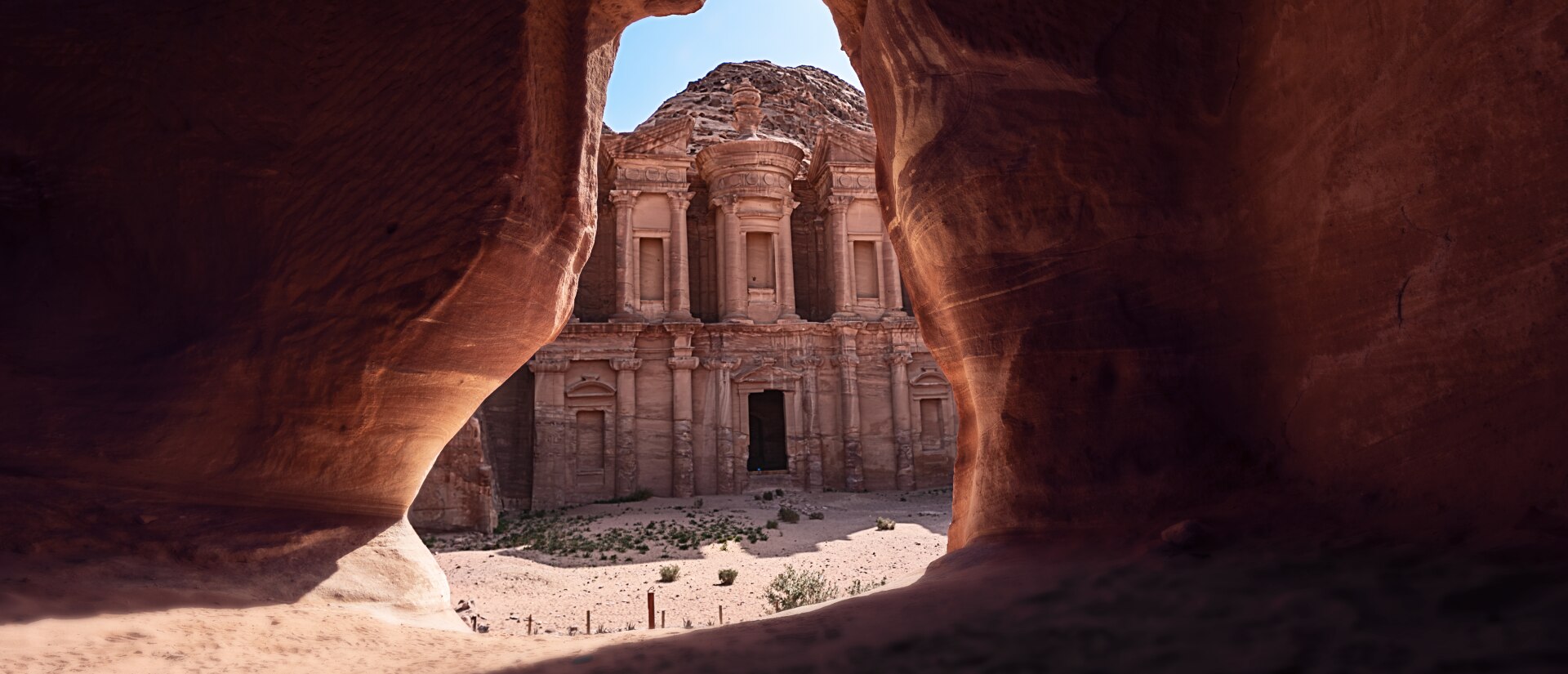 View from inside a cave in Jordan looking out at Petra, a structure partially carved into the rock and surrounded by mountains.