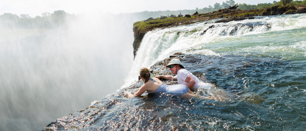 A man and woman laying in the shallow water at the edge of Victoria Falls in Zimbabwe.