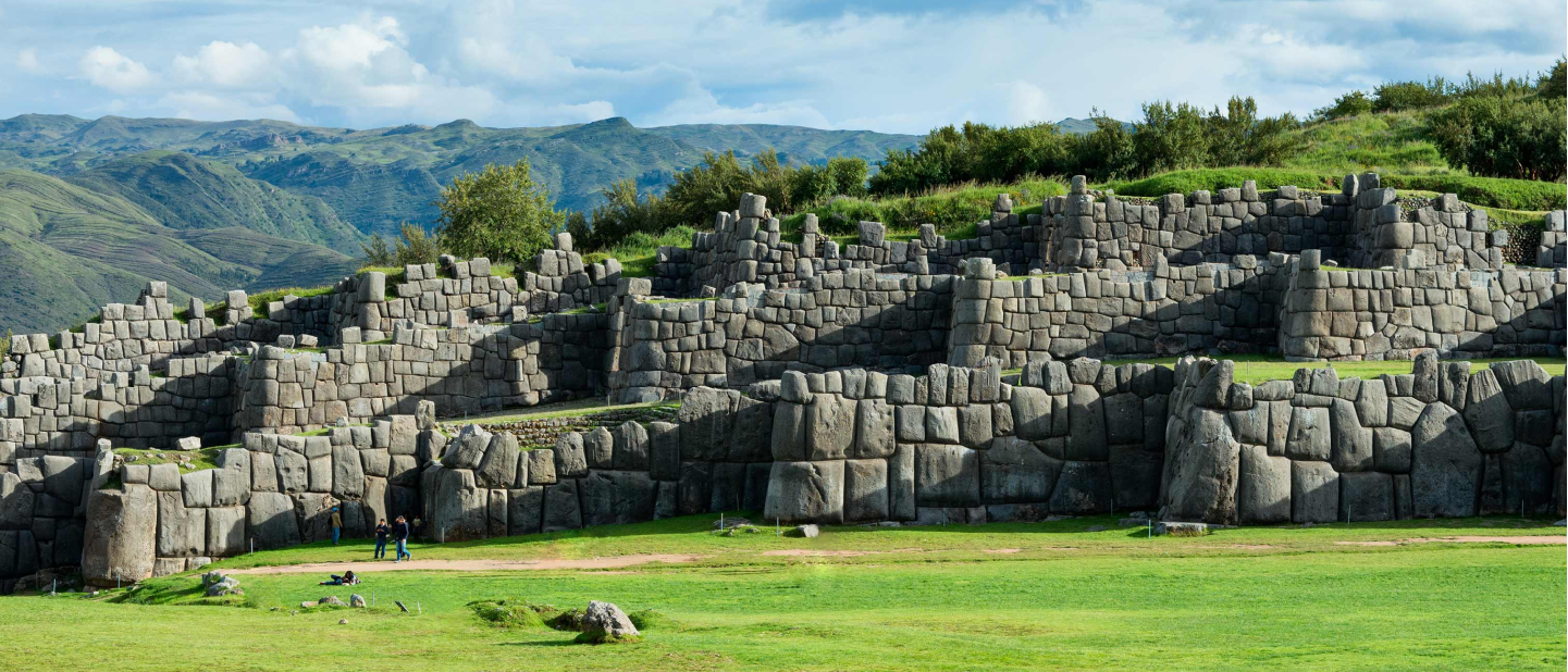 Ground view of the Sacsayhuaman, a monumunt made of stone built into the hillside. Plush, green grass lays in front.