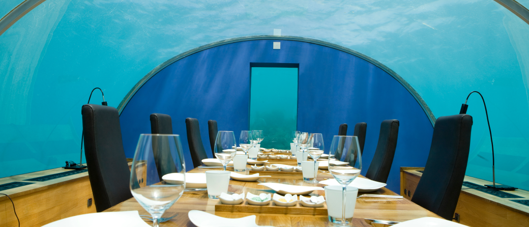 A set dining room table located in an underwater hotel. The walls and ceiling are windows which look out into the ocean.