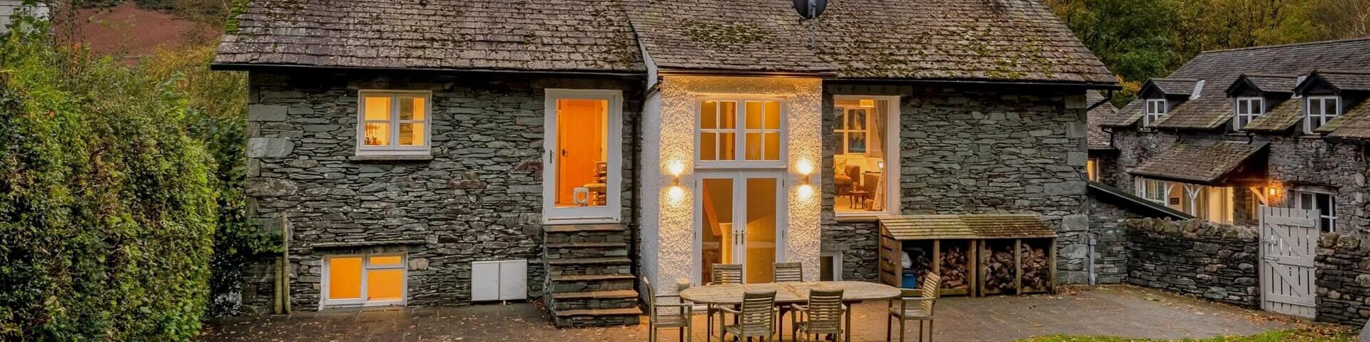 A cosy stone cottage at dusk surrounded by trees, with light shining through the windows.