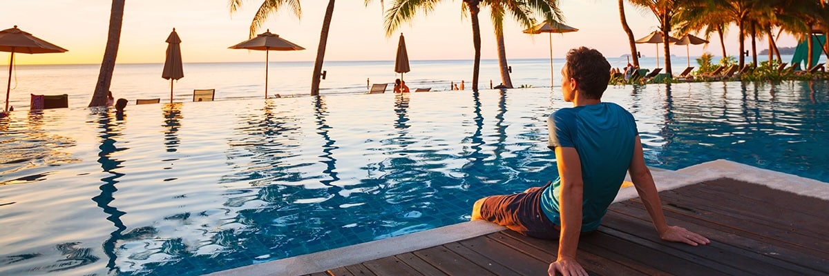 A man sits in an infinity pool and faces the setting sun.