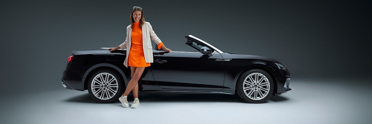 A woman in an orange dress leans up against a black convertible rental car. Sixt Car Rentals.