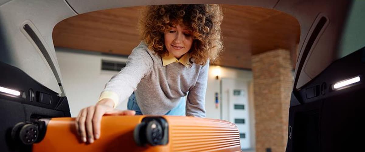 A woman reaches for her orange luggage from the back of a car.