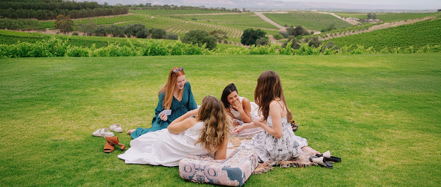 Two people enjoy a relaxing moment in a lush garden, surrounded by vibrant flowers, as one offers something while the other holds a glass of white wine.