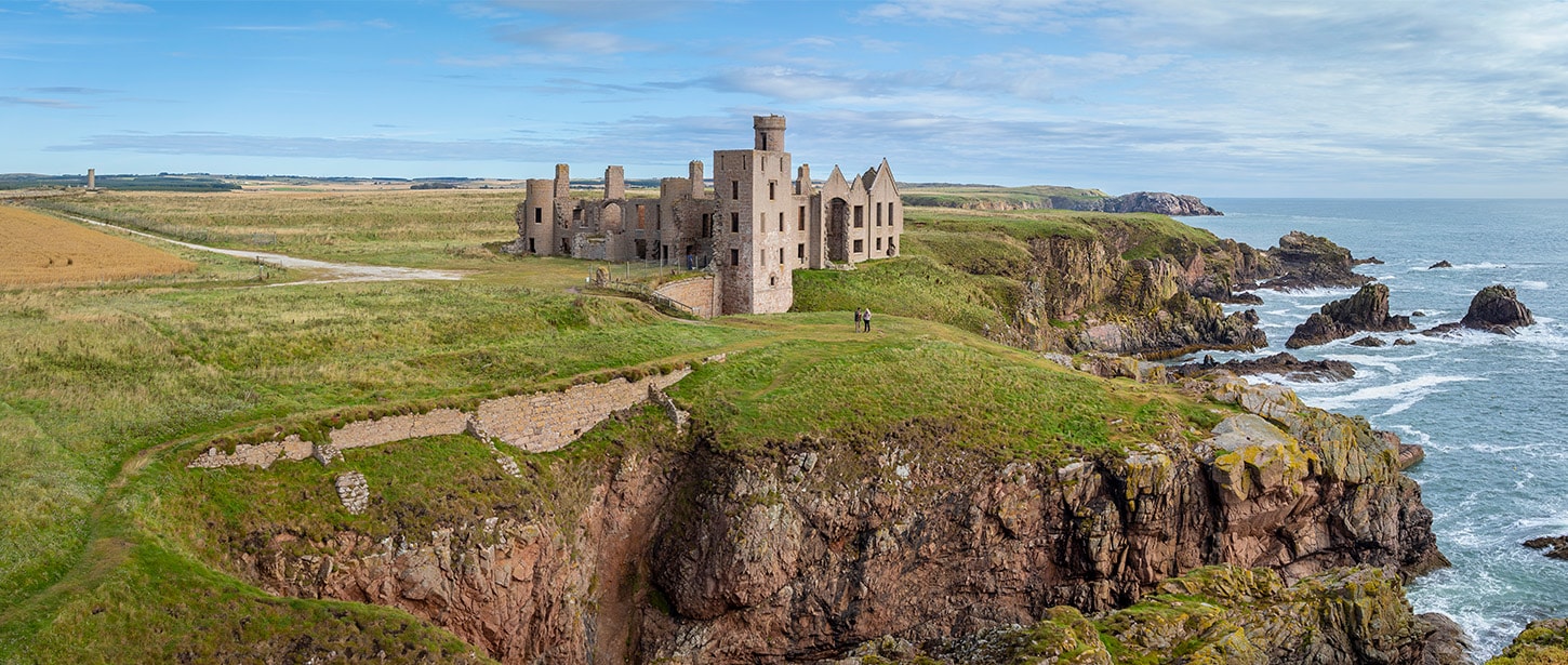 Ruined castle on a grassy cliff above the ocean with waves crashing below.