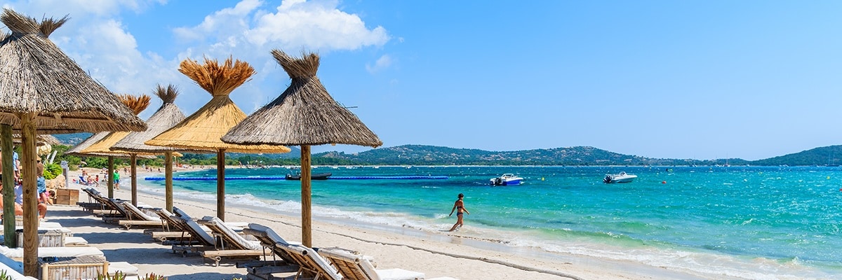 A scenic beach with blue water, boats, straw-roofed umbrellas and lounge chairs.
