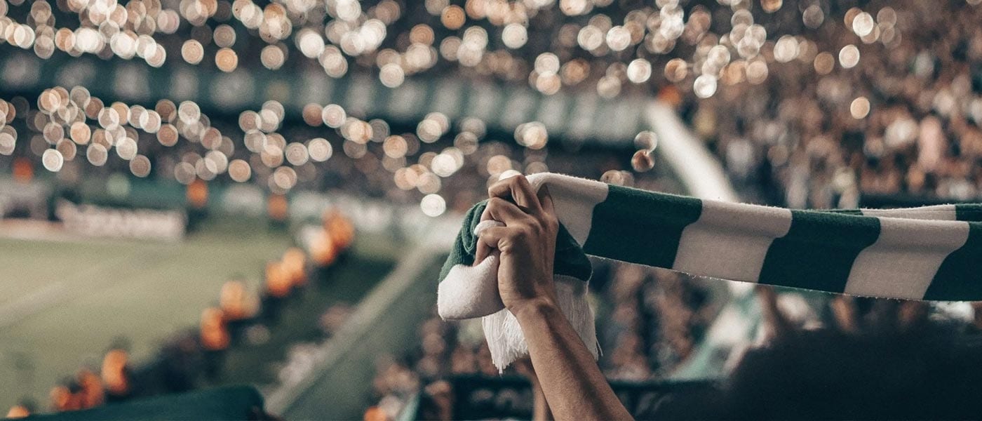 Close-up of hands holding a green and white striped scarf above their head in a crowded soccer stadium.