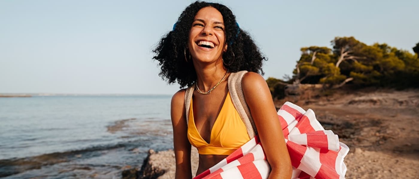 A smiling woman on the beach carrying a red and white beach umbrella.