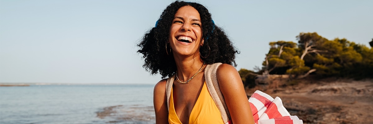 A smiling woman on the beach carrying a red and white beach umbrella.