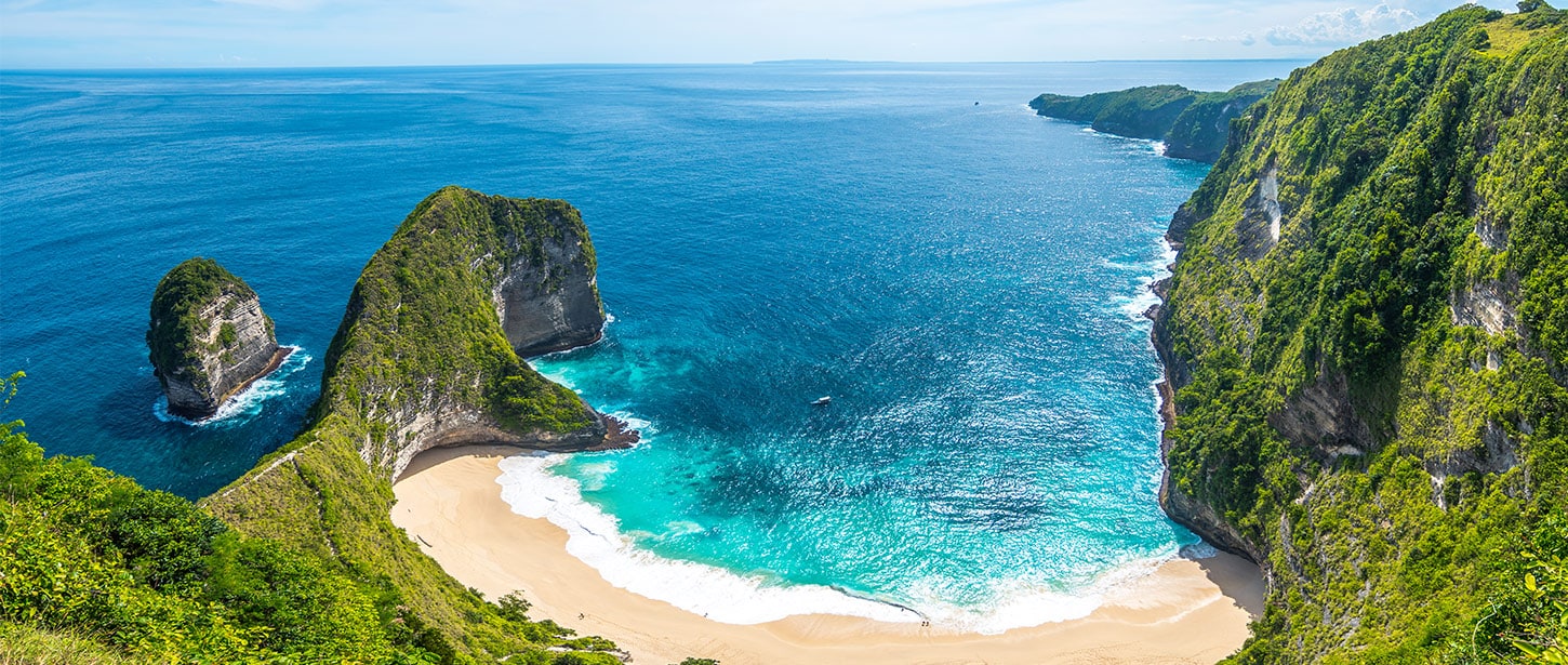 Aerial view of a secluded tropical beach with golden sand, turquoise water, and dramatic green cliffs rising above the ocean.