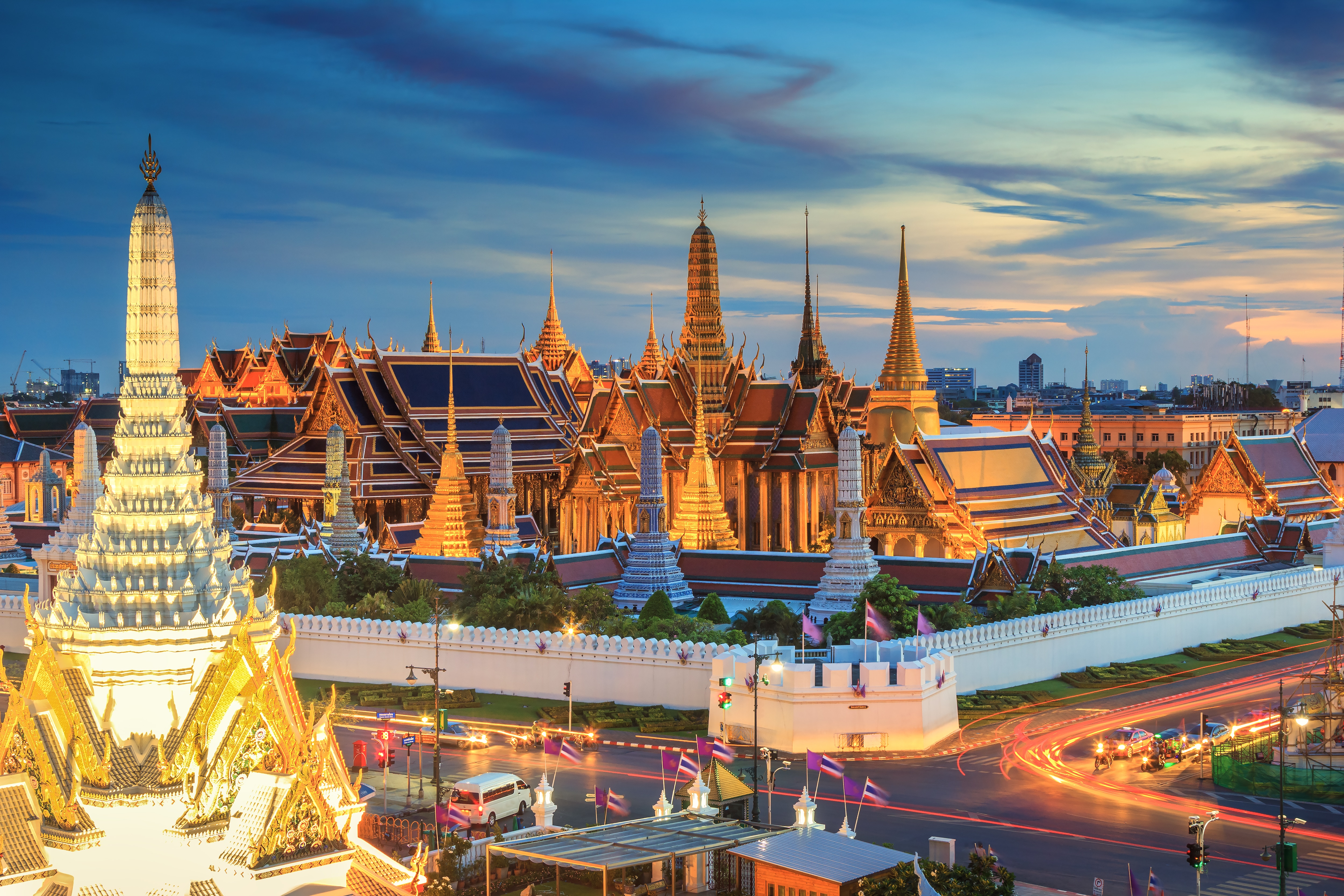 Vue aérienne du Grand Palais illuminé et du complexe du temple Wat Phra Kaew à Bangkok, en Thaïlande, au crépuscule, avec des flèches dorées resplendissantes se détachant sur un ciel bleu dramatique.