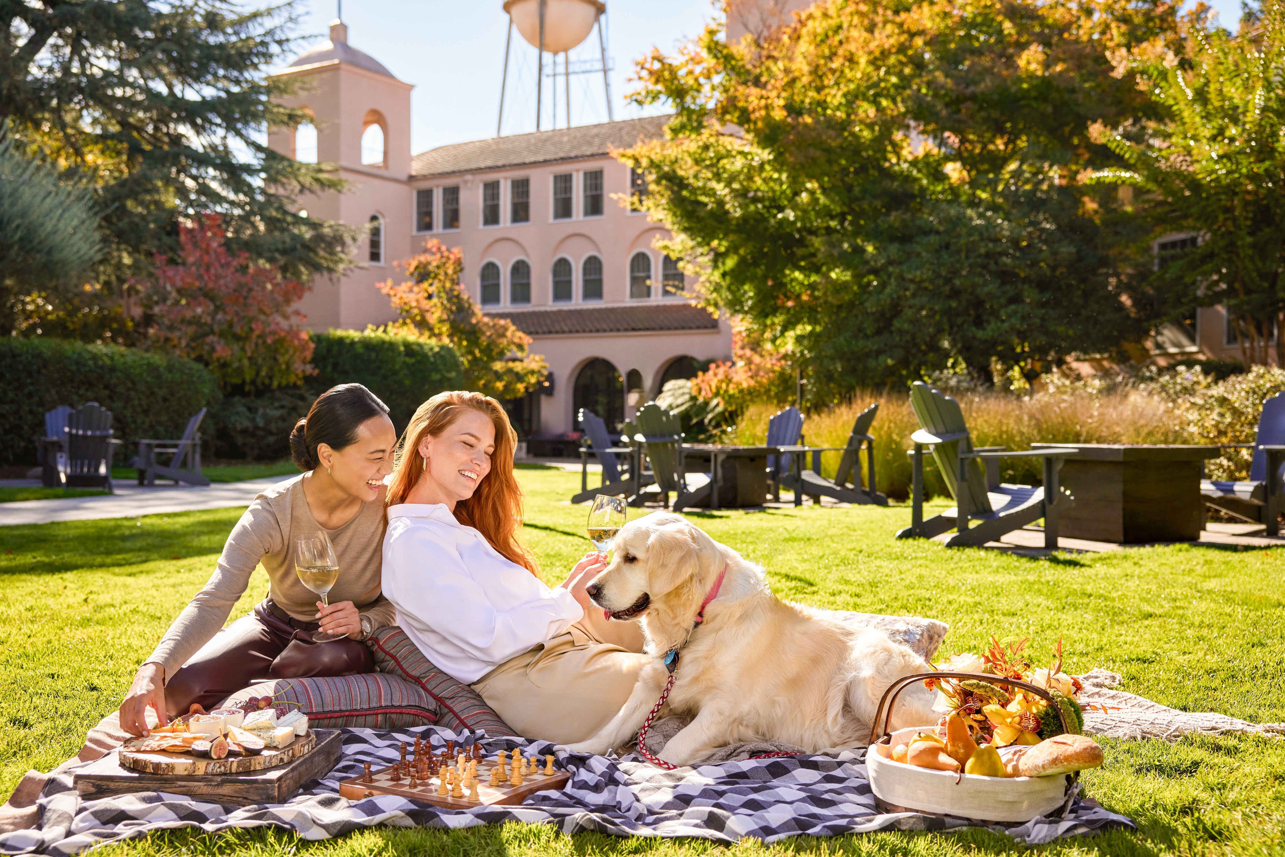 Deux femmes se détendent sur une couverture de pique-nique sur la pelouse ensoleillée d'un hôtel avec un golden retriever, des verres de vin blanc, un plateau de charcuterie et un jeu d'échecs, avec un charmant bâtiment d'hôtel en arrière-plan.