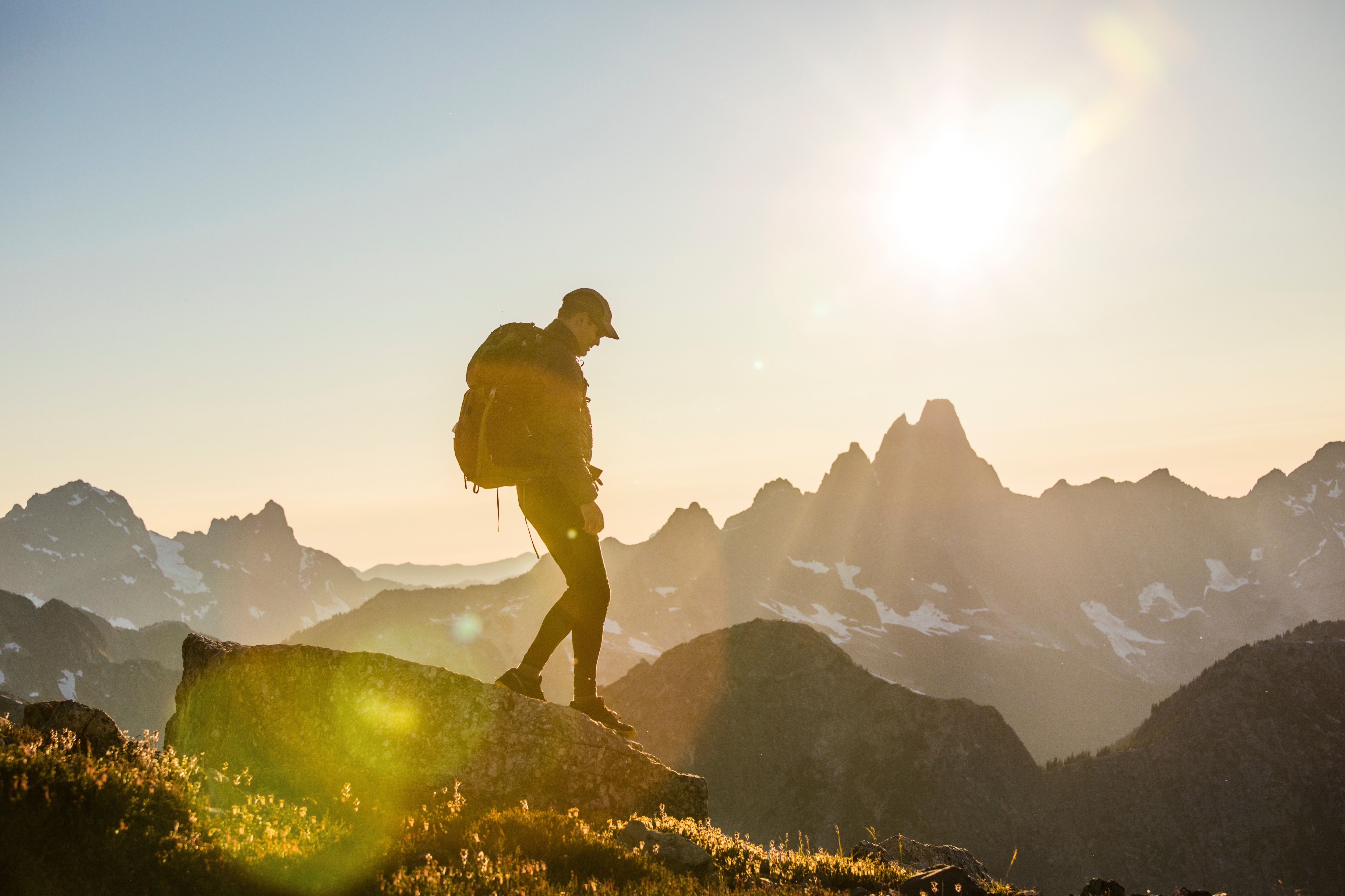 A hiker silhouetted against a golden sunset stands on a rocky mountain summit, wearing a backpack and cap, with dramatic snow-capped mountain peaks and a glowing sun in the background.