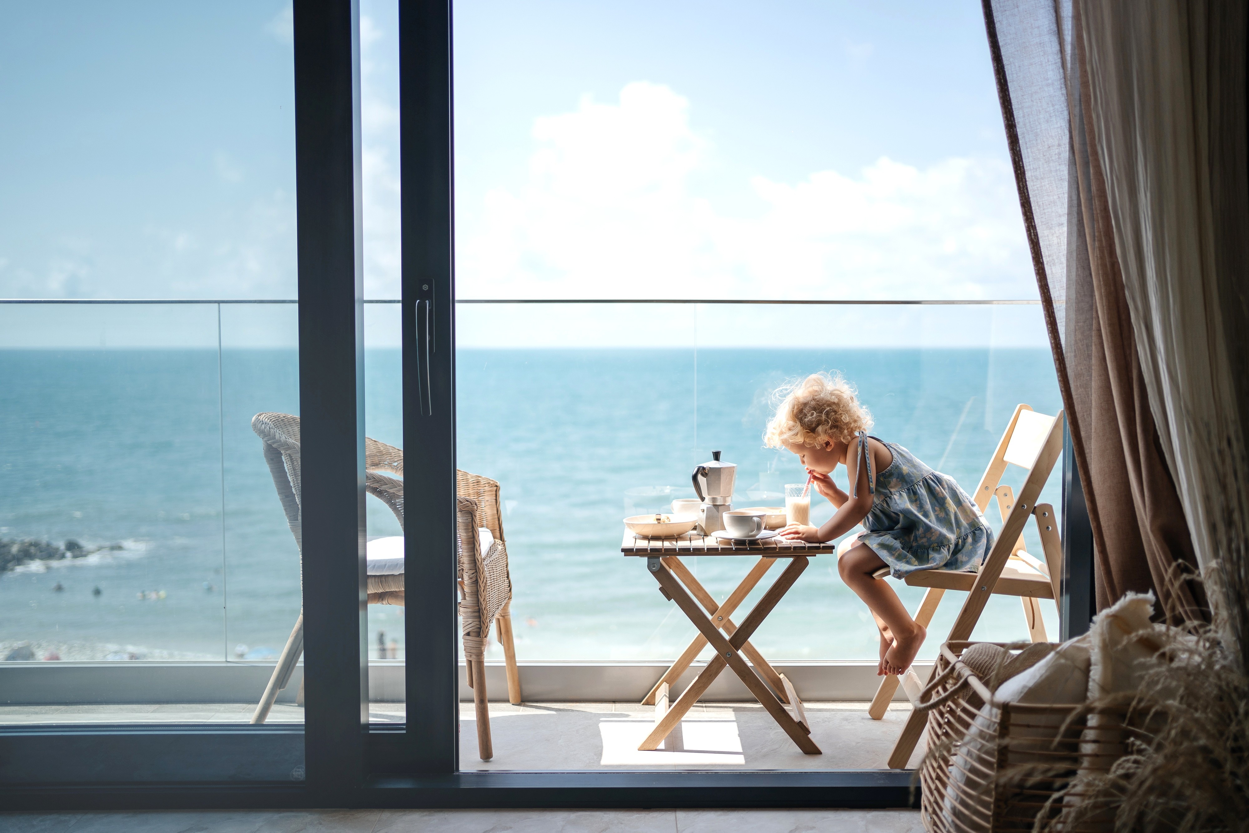 Ett litet barn med lockigt hår sitter på en balkong med utsikt över havet och äter frukost vid ett litet träbord på en solig dag.