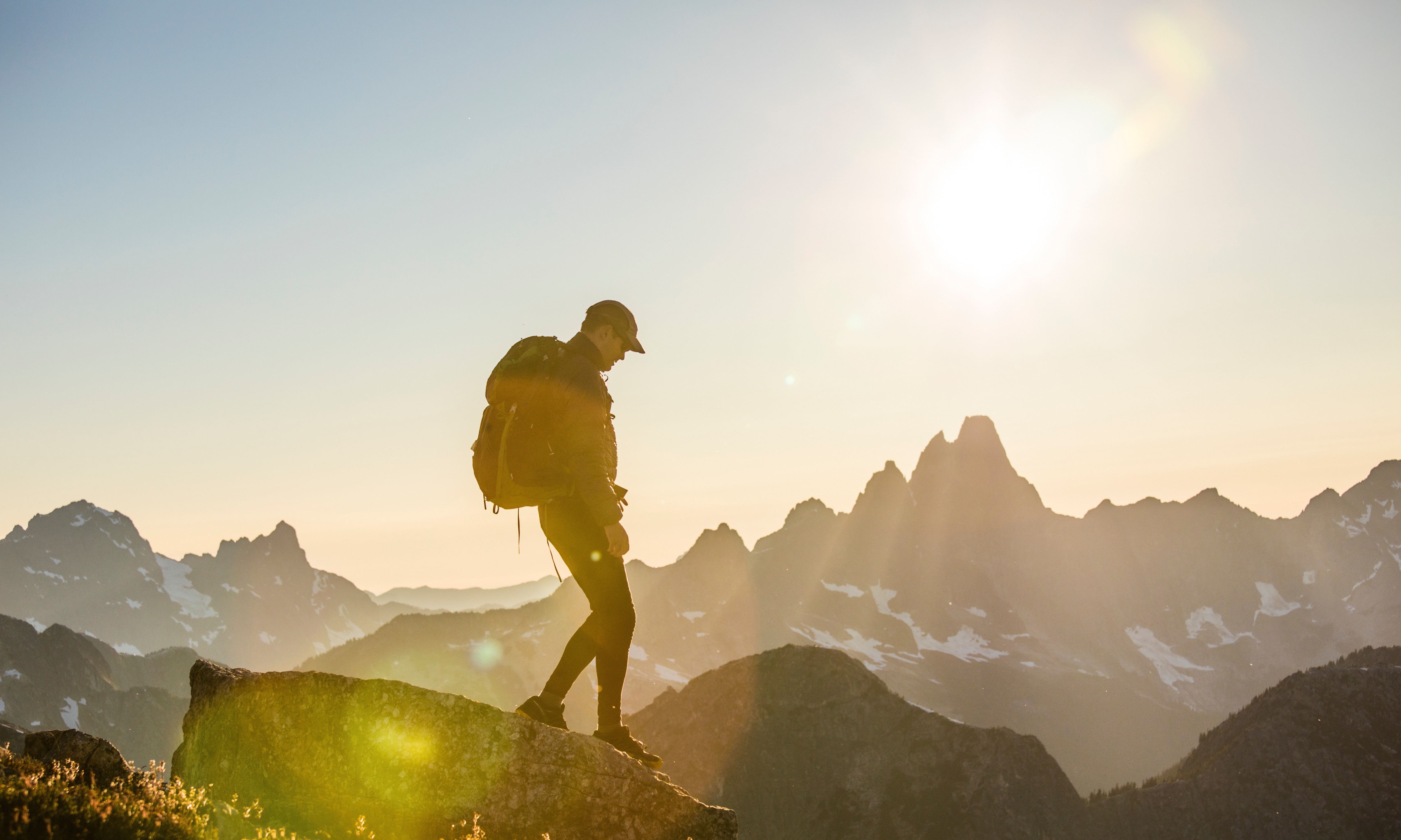 A hiker silhouetted against a golden sunset stands on a rocky mountain summit, wearing a backpack and cap, with dramatic snow-capped mountain peaks and a glowing sun in the background.