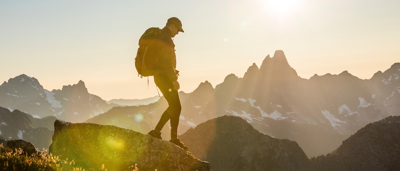 A hiker silhouetted against a golden sunset stands on a rocky mountain summit, wearing a backpack and cap, with dramatic snow-capped mountain peaks and a glowing sun in the background.