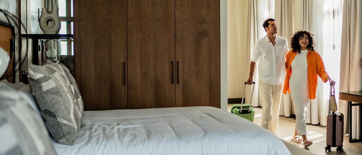 A couple enters a hotel room with rolling luggage.