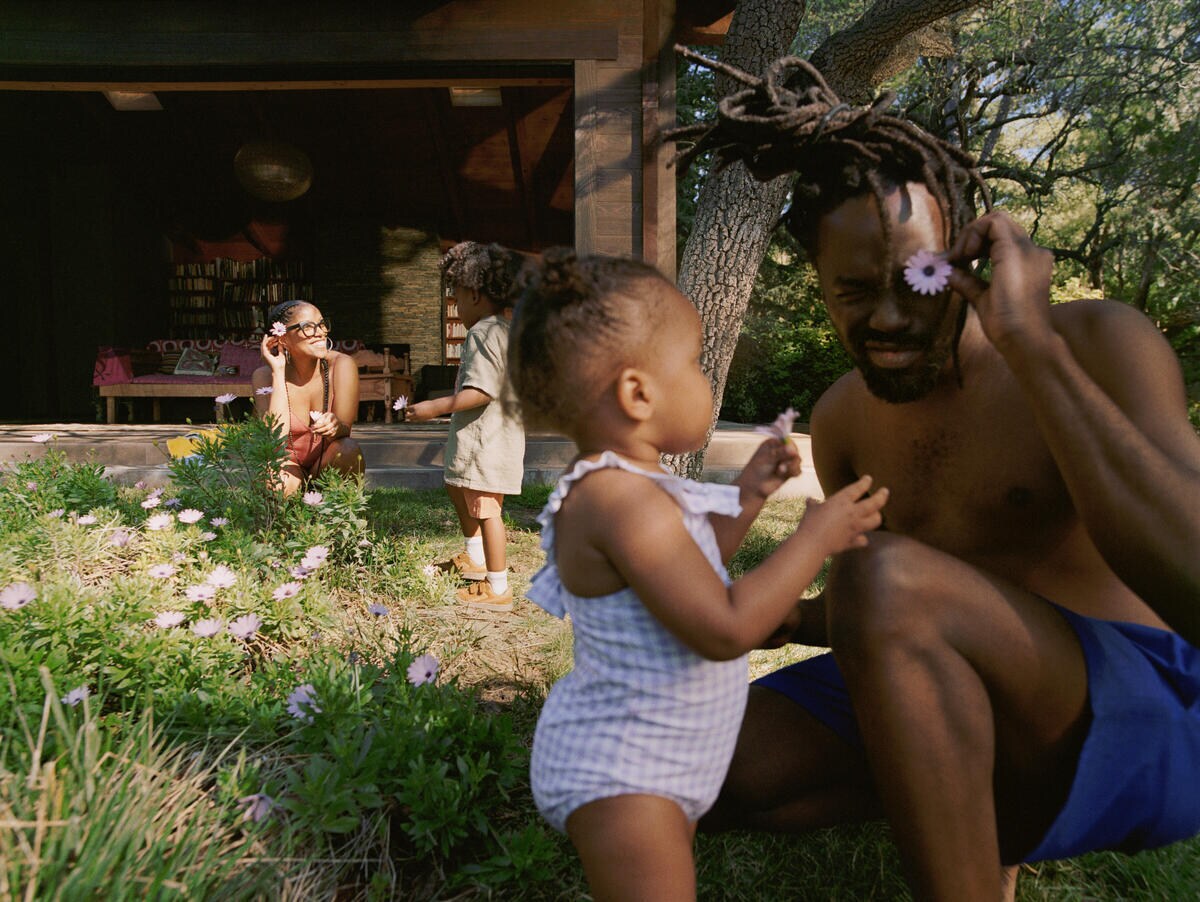 In the foreground a man and baby girl play with flowers they've picked from a flower patch beside them. In the background a smiling woman and a little boy are also holding flowers.