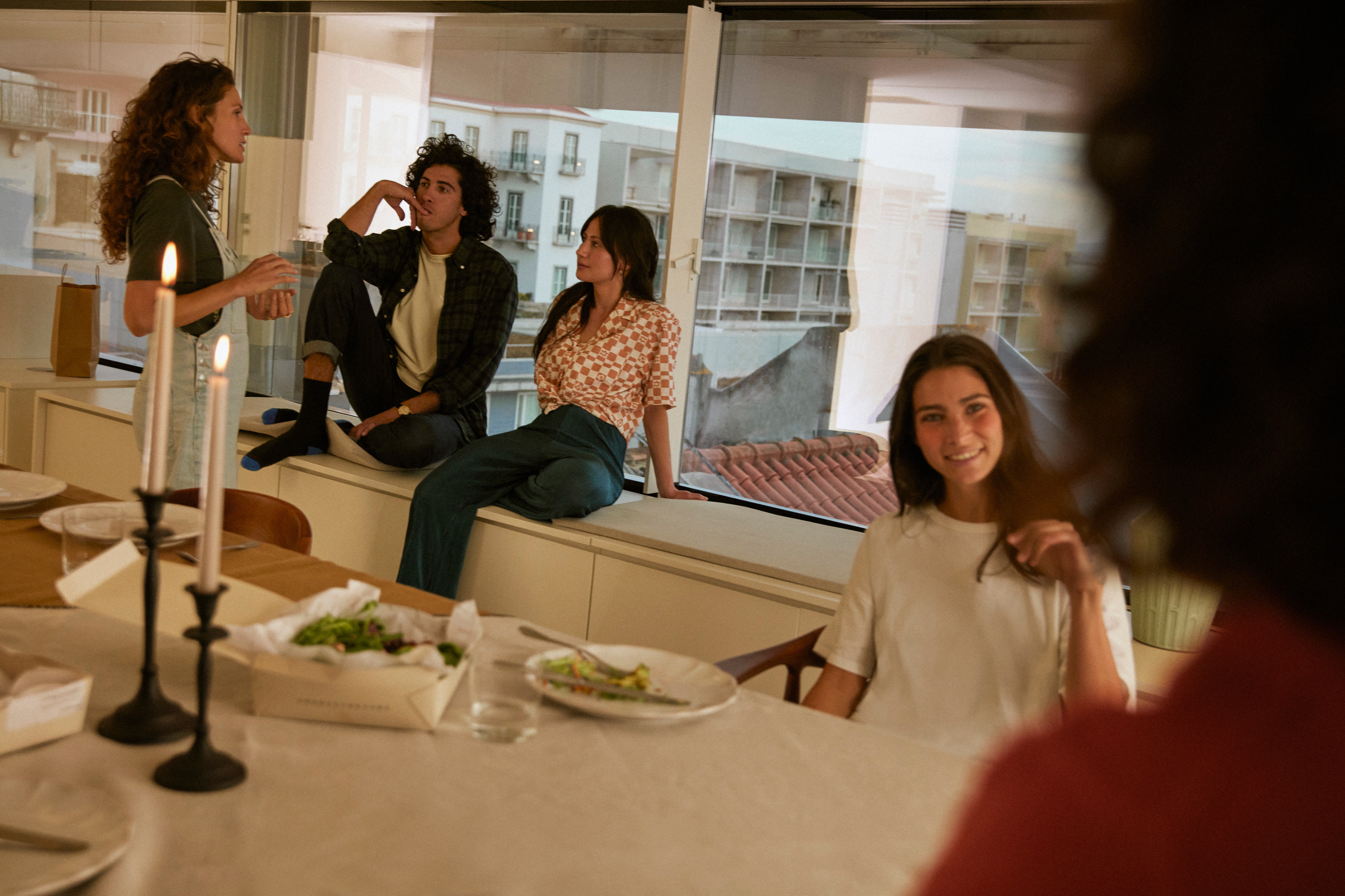 A group of friends share a meal as they sit at a counter and along a window sill in a bright kitchen.