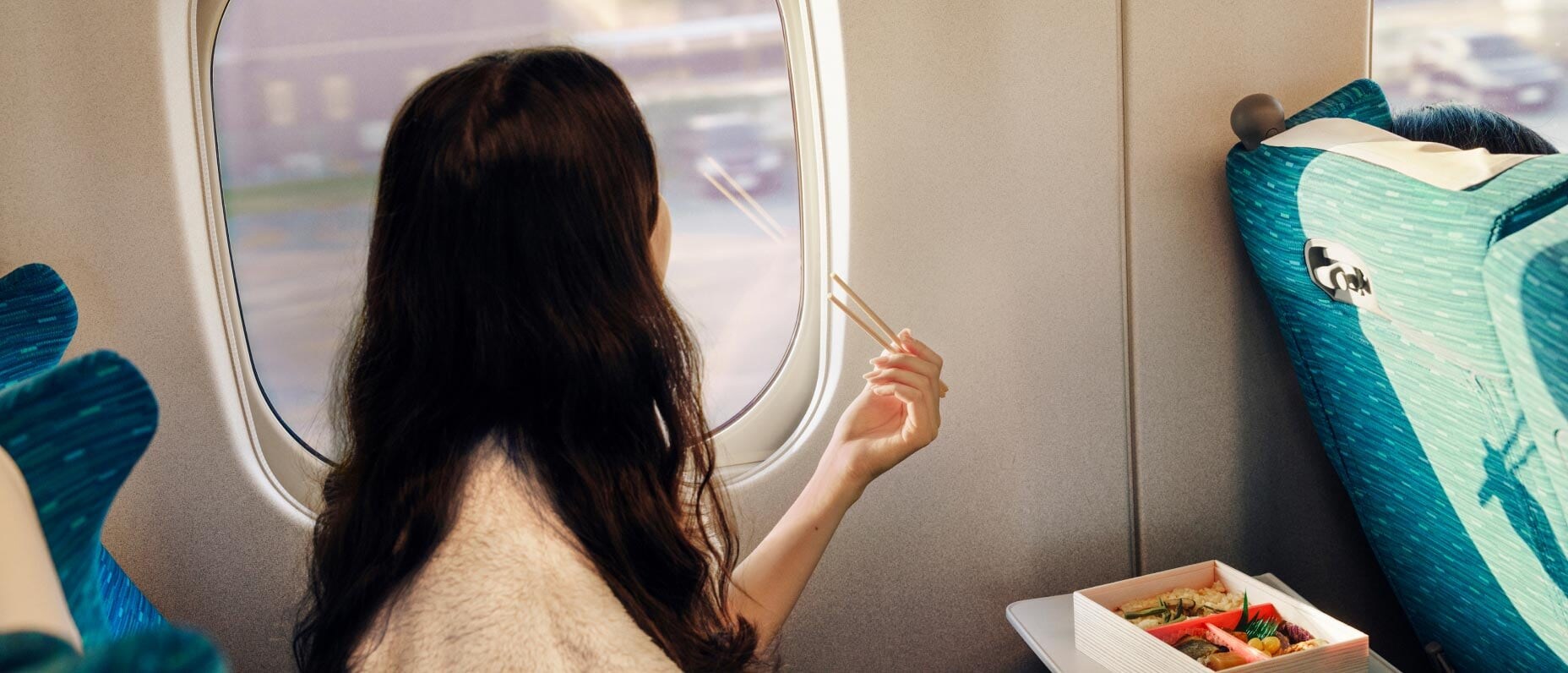 A woman wearing a white sweater sitting on a bullet train looking out the window holding chopsticks and eating sushi.