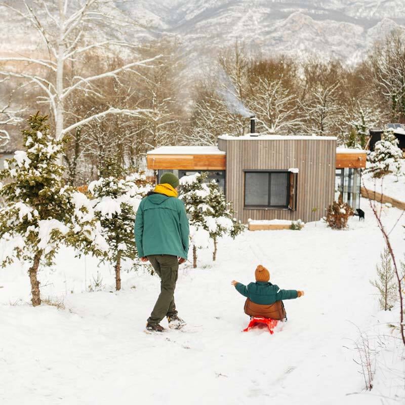 De la fumée s’élève d’une cabane moderne nichée dans un paysage boisé et enneigé.  Sur un chemin qui descend vers l’hébergement, un adulte marche aux côtés d’un enfant sur une luge, tous deux bien emmitouflés pour se protéger du froid.
