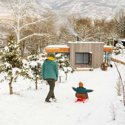 De la fumée s’élève d’une cabane moderne nichée dans un paysage boisé et enneigé.  Sur un chemin qui descend vers l’hébergement, un adulte marche aux côtés d’un enfant sur une luge, tous deux bien emmitouflés pour se protéger du froid.