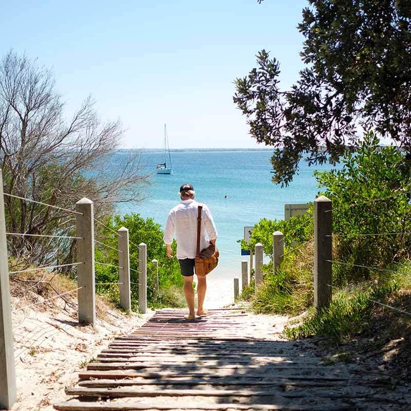 A man walks barefoot down a rustic path to a white-sand beach in the sunshine, with a yacht moored in the turquoise sea beyond. He wears a white linen shirt, dark shorts and a dark baseball cap, and carries a leather bag.