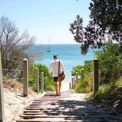 A man walks barefoot down a rustic path to a white-sand beach in the sunshine, with a yacht moored in the turquoise sea beyond. He wears a white linen shirt, dark shorts and a dark baseball cap, and carries a leather bag.