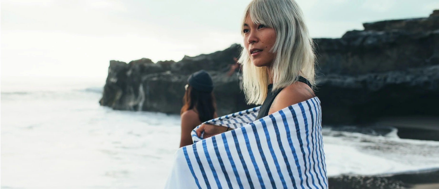 A woman with blonde hair stands on a beach, wrapped in a striped towel, gazing thoughtfully at the ocean. A rocky shoreline is visible in the background.