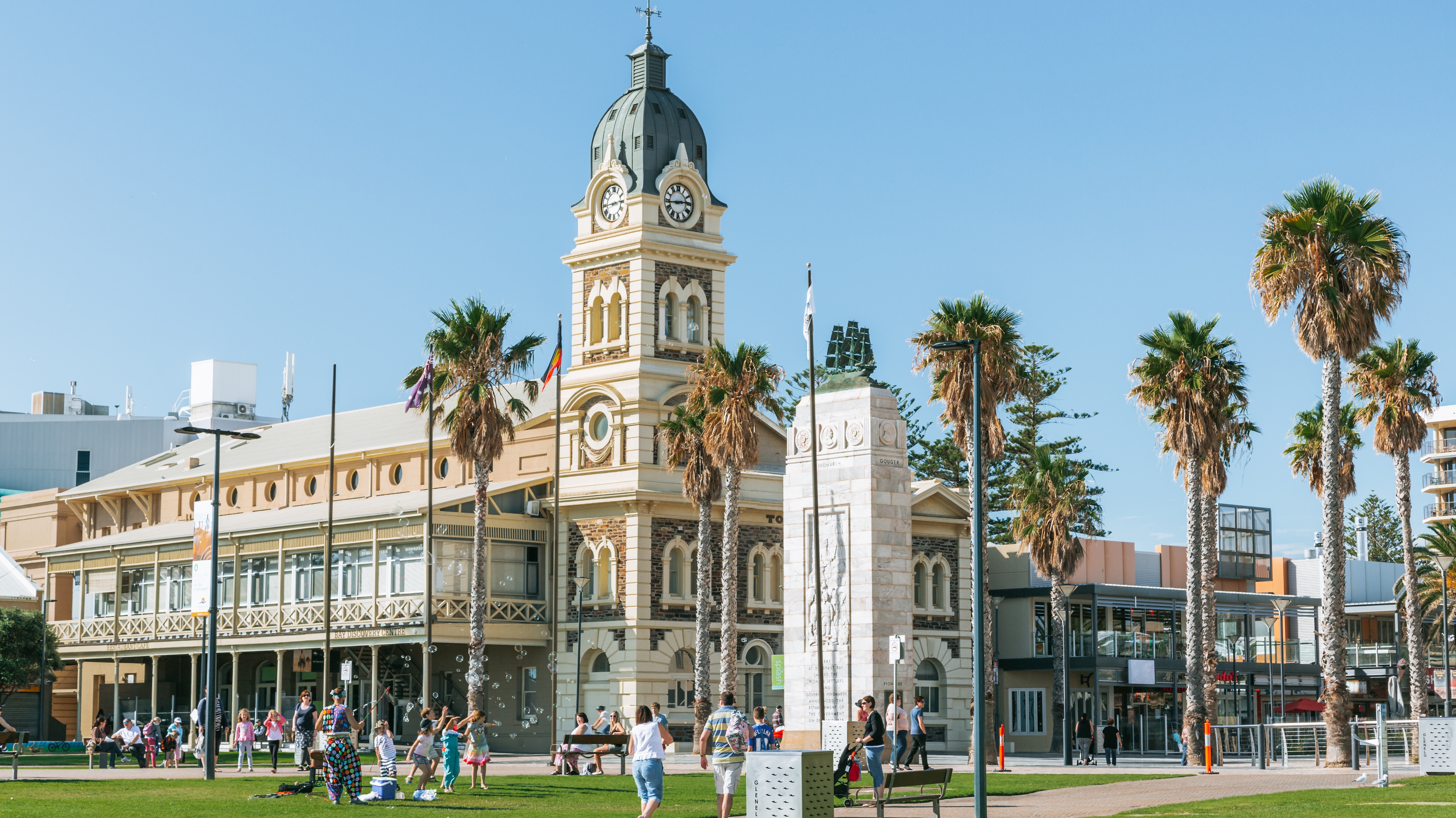 Glenelg Beach in Adelaide, South Australia offers a vibrant atmosphere with palm trees, historic architecture, and families enjoying leisure activities in bright sunlight