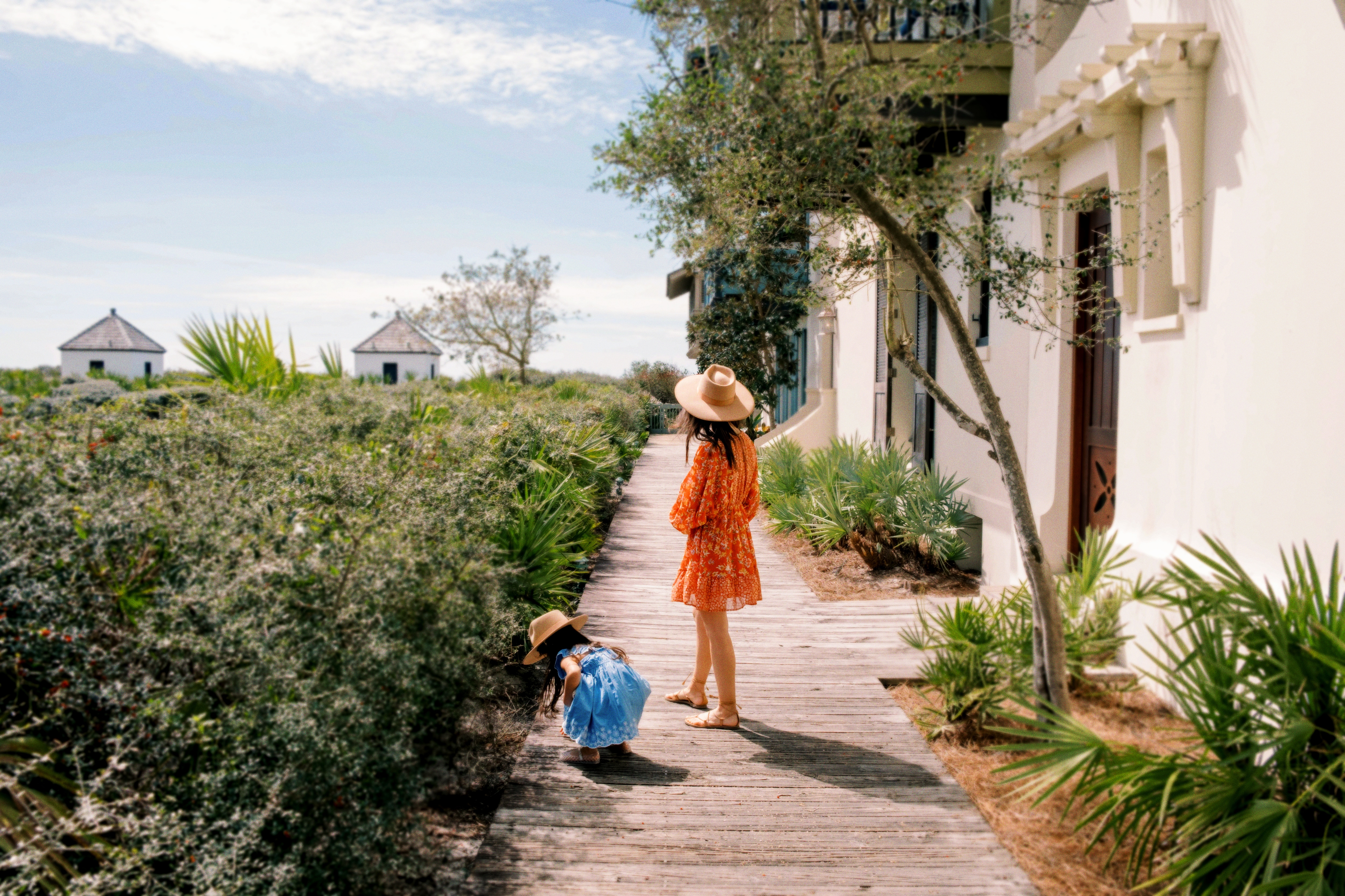 A woman and a young girl wearing hats and summer clothing walk along a wooden boardwalk surrounded by lush greenery and white buildings near the coast.