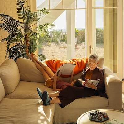 Escapades littéraires à la plage, un jeune garçon et une femme lisant des livres sur un canapé beige dans la salle de séjour d’une maison de plage baignée de soleil, mettant en valeur une propriété de vacances côtière décontractée.