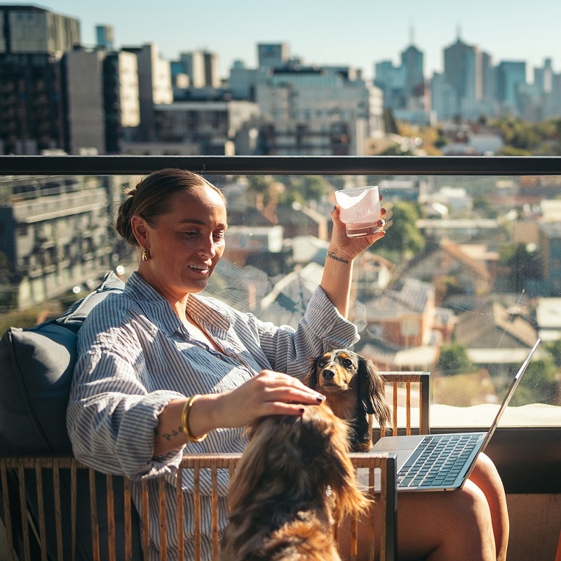 Two women sit on a balcony with drinks and a dog, overlooking a sunlit cityscape.