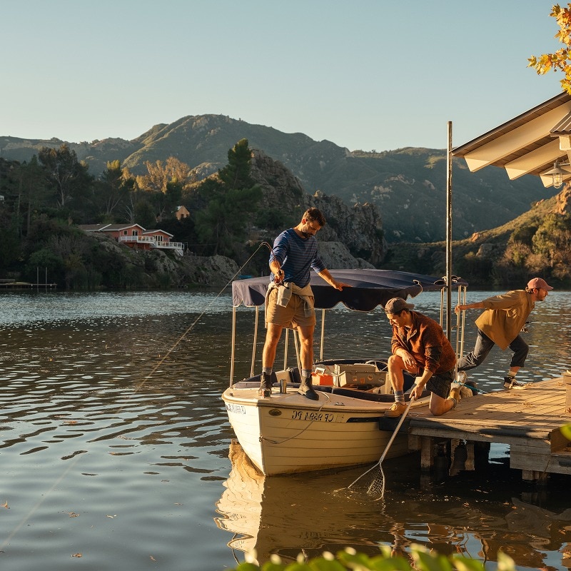 Three men on a wooden dock on a lake, surrounded by a hilly, wooded landscape on a sunny day.