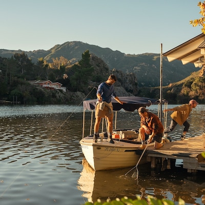 Three men on a wooden dock on a lake, surrounded by a hilly, wooded landscape on a sunny day.