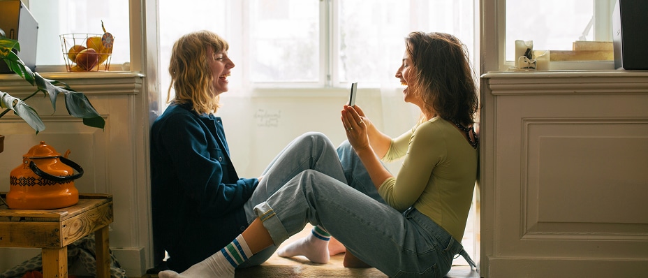 Two women sit smiling on a sunlit floor by the window, sharing a relaxed moment indoors. 