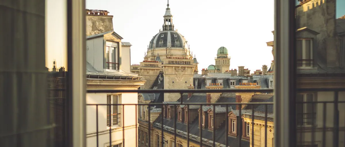 Vue depuis une fenêtre ouverte d’un balcon parisien, donnant sur les toits des immeubles classiques et le dôme de la chapelle de la Sorbonne. 